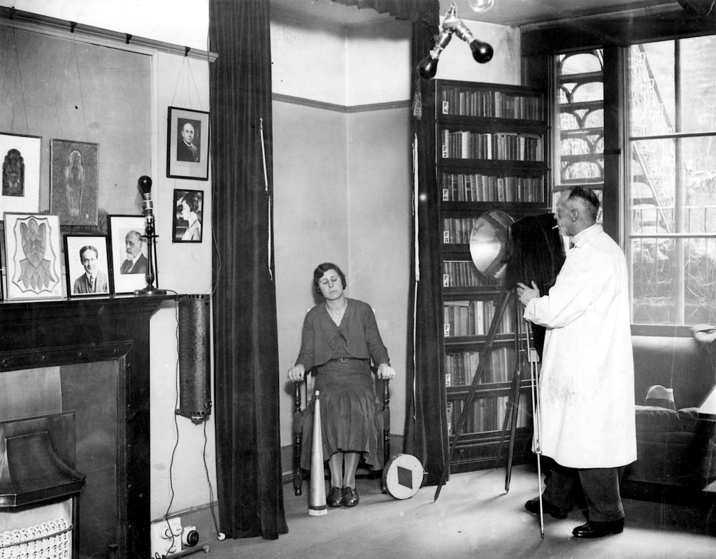Le chercheur londonien Harry Price et sa secrétaire Ethel Beenham en train de tenter de reproduire des photos de spirites sur lesquelles seraient imprimées des apparitions spectrales. A gauche sur la cheminée, une photo de Harry Houdini, le célèbre magicien qui lui aussi traqua les faux spirites.